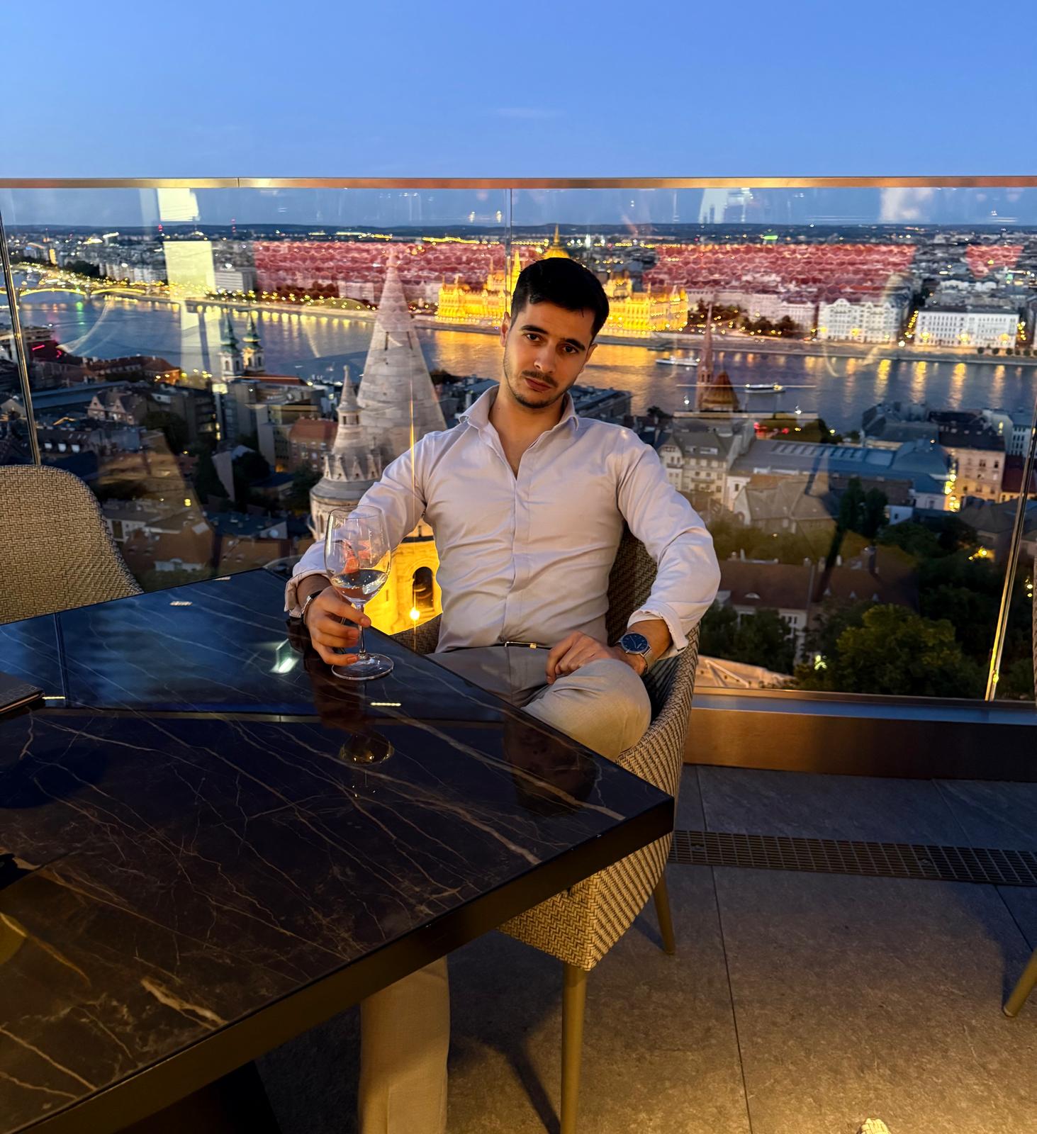 Csongor Osz seated on a terrace overlooking a river and city skyline at dusk